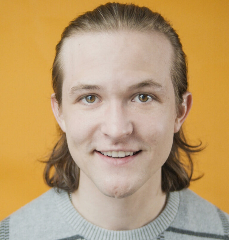 Actor Danny Irizarry wearing a gray sweater in front of an orange background.