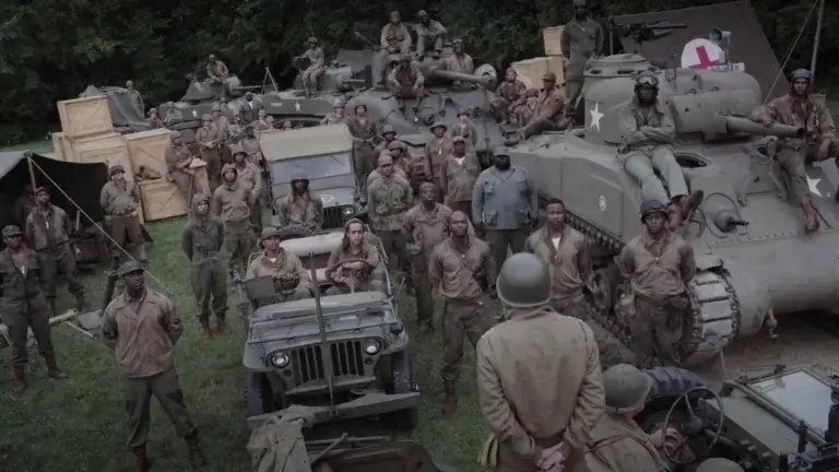 A group of military men in army fatigues at a base surrounded by army cars and a tank awaiting instructions.