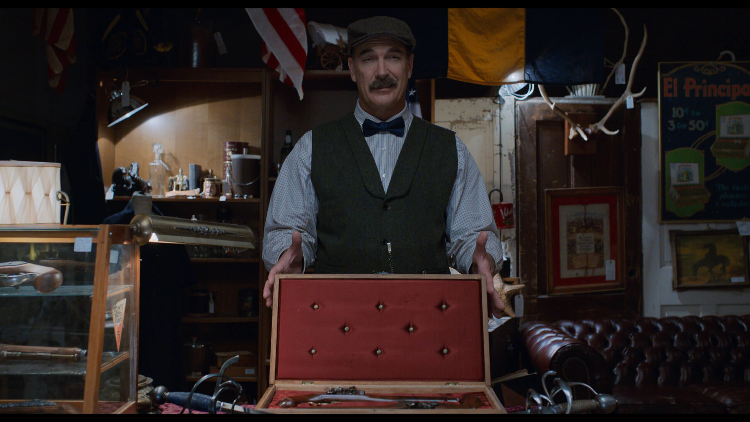 Patrick Warburton in old timey clothes showing off old dueling pistols in an antique shop.
