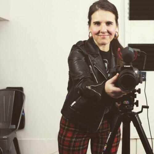 Mia Cusumano in plaid pants and a leather jacket behind a video camera.
