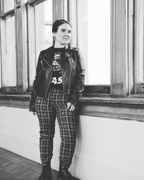 Black and white photo of Mia Cusumano in plaid pants and a leather jacket inside.