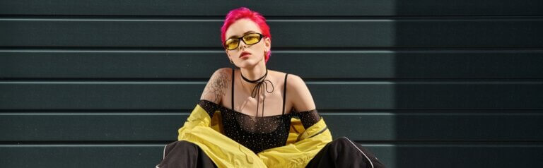 Hip young woman with tattoos piercings and pink hair sitting in front of a black garage door.
