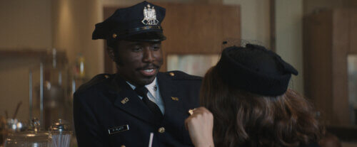 Y’lan Noel in a police uniform smiling inside a diner talking to a woman in a mourner's hat.