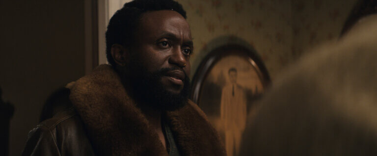 Byron Bowers inside a 1950's house wearing a brown jacket looking concerned.