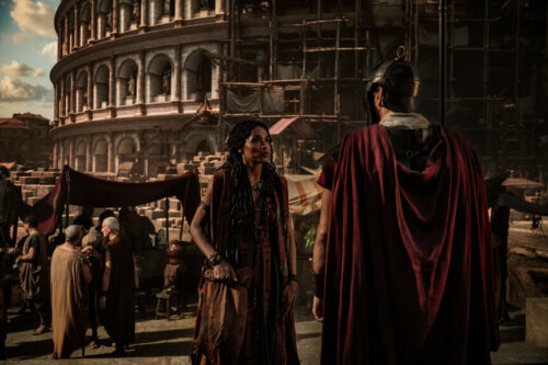 Sara Martins in Roman robes speaking with a Roman soldier outside a colosseum in Peacock's 'For Those About to Die.'