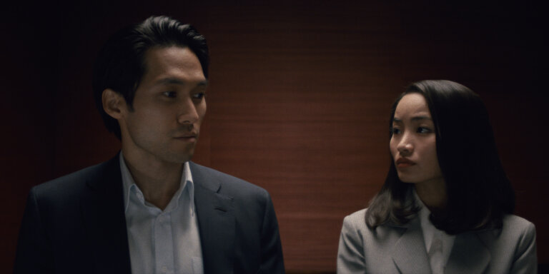 Jin Ha and Anna Sawai in "Pachinko" wearing business attire and looking nervous in an elevator.