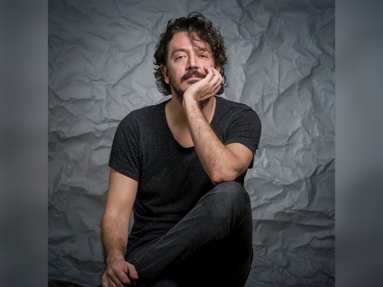 Director/writer Tolga Karaçelik in a black shirt and jeans looking stoic by a gray wall.