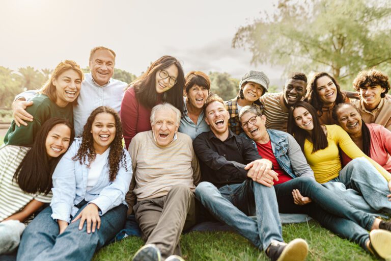 Diverse group of multigenerational people smiling and sitting on grass in a public park.