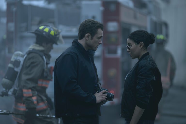 Taron Egerton and Jurnee Smollett in jackets outside investigating a fire has a firefighter walks by.