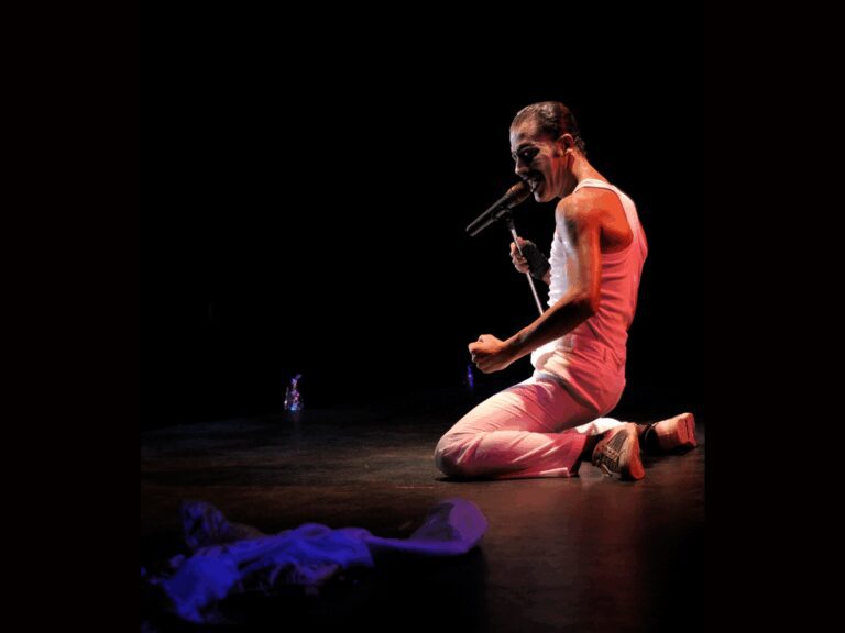 Freddie Mercury lookalike singer performing onstage , on his knees with a fist clenched.